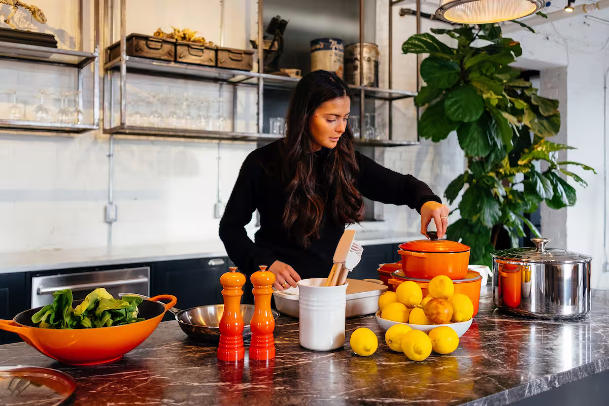 Una mujer de cabello oscuro en una cocina moderna con encimera de mármol está cerca de ollas y utensilios de cocina naranjas y plateados, así como un tazón de limones. Se ven ingredientes frescos como hojas verdes en un wok naranja.